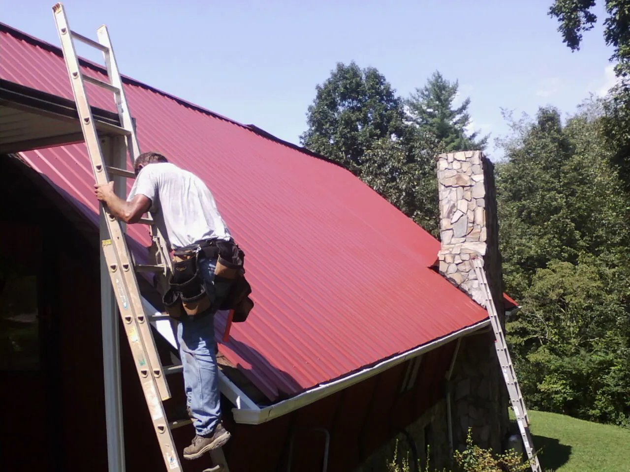 Roofer on a ladder beside a red metal roof and stone chimney — Soffit & Fascia Repair by Roofing Brothers in Bradfordsville